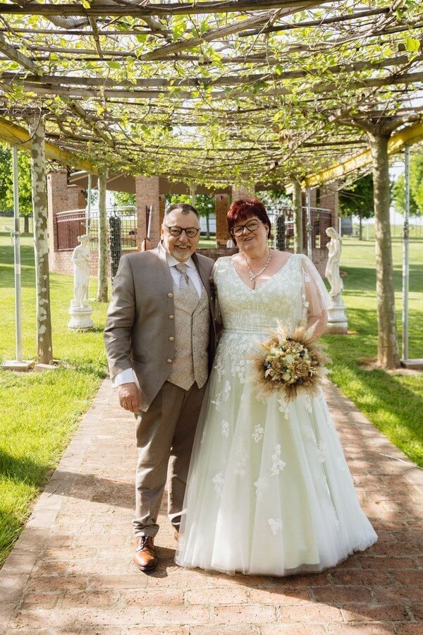 Curvy Braut in salbeigrünem Jarice-Brautkleid mit kleiner Ärmellösung und langer Schleppe, Bräutigam in hellbraunem Tziacco-Stehkragenanzug mit Weste – Hochzeitsmode Übergröße bei Biancas Brautmoden