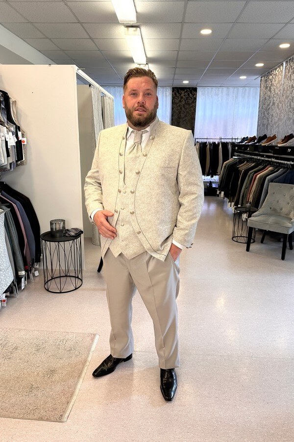 Bräutigam in großer Größe trägt eleganten Tziacco-Gehrock mit Stehkragen, gemusterter Jacke und Plastron – Hochzeitsanzug in Beige bei Biancas Brautmoden
