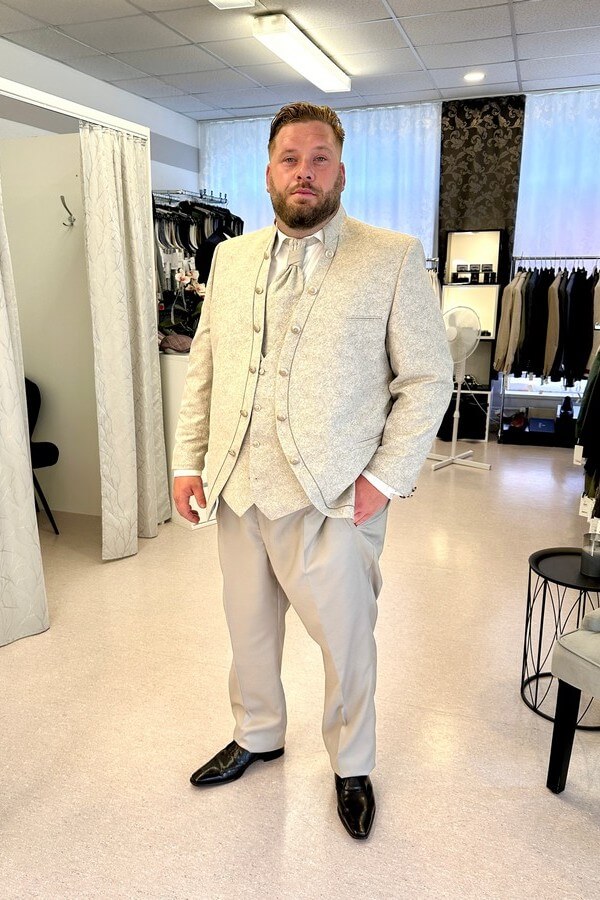Bräutigam in großer Größe trägt eleganten Tziacco-Gehrock mit Stehkragen, gemusterter Jacke und Plastron – Hochzeitsanzug in Beige bei Biancas Brautmoden