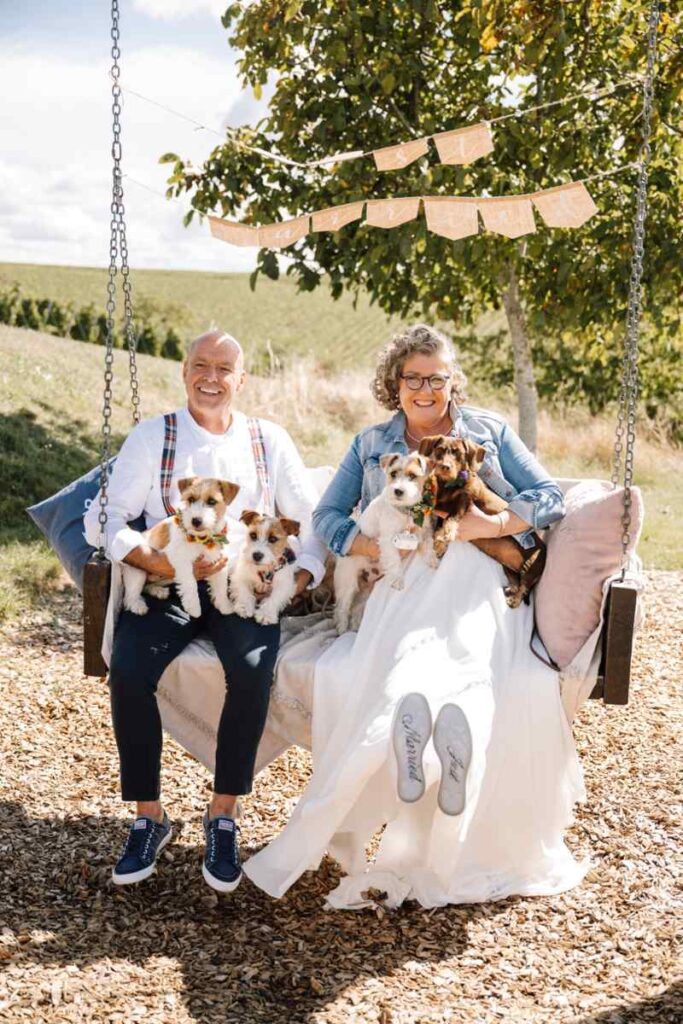 Curvy Braut Ü50 mit Jeansjacke und Bräutigam mit Hosenträgern sitzen mit ihren Hunden auf einer Schaukel – Bianca’s Brautmoden Curvybrautmode Rheinland-Pfalz.