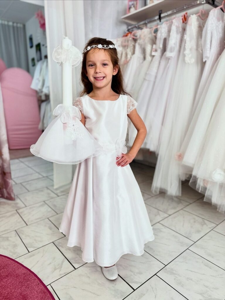 Kommunionkleid Auswahl. Kommunionkleid in Weiß mit Satin, Spitzenärmeln und Taillenblüte – klassisches Kommunionoutfit mit Kerze, erhältlich bei Bianca’s Brautmoden Bad Münster.