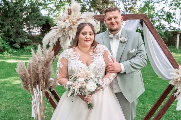 Brautpaar in großer Größe an ihrer Hochzeit. Er im salbeigrünen Anzug, sie in einem Brautkleid Plussize in A-Linie mit langen Ärmeln.