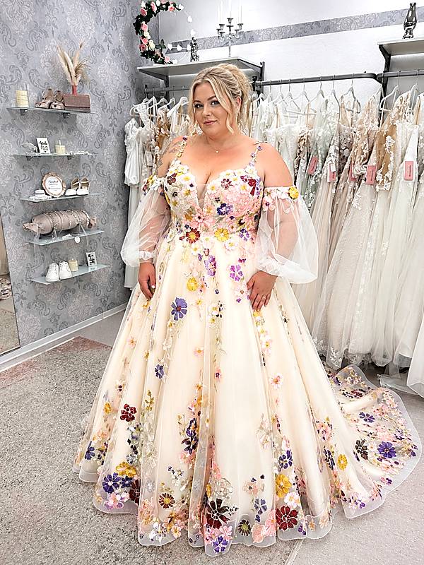 Curvybraut im romantischen Brautkleid mit farbigen 3D-Blumenstickereien, transparenten Puffärmeln und weiter A-Linie in einem Brautmodengeschäft.