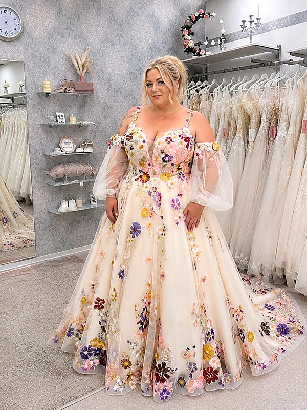 Curvybraut im romantischen Brautkleid mit farbigen 3D-Blumenstickereien, transparenten Puffärmeln und weiter A-Linie in einem Brautmodengeschäft.