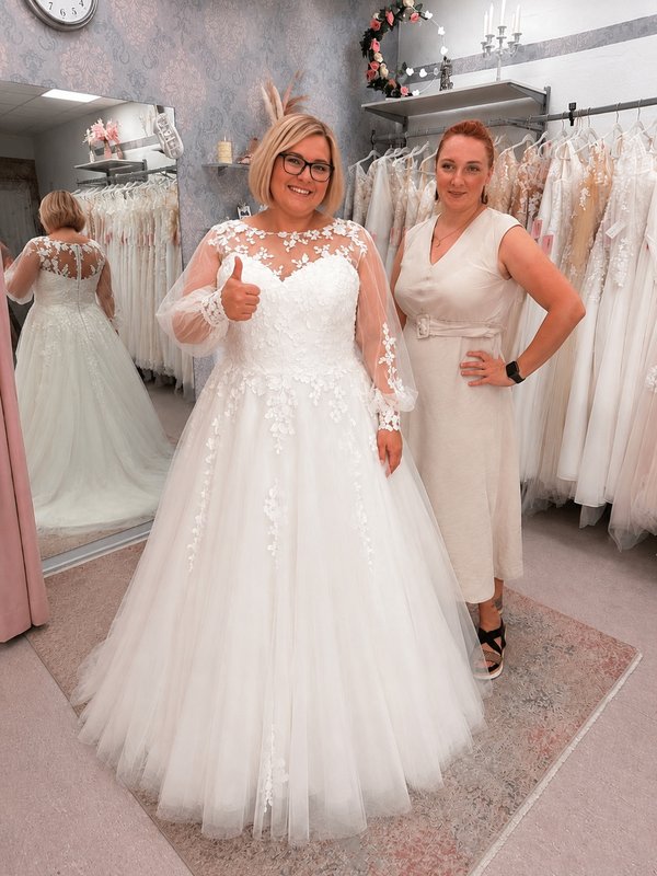 Glückliche Curvy Braut mit Brautberaterin nach erfolgreicher Anprobe eines Brautkleids mit langen Spitzenärmeln bei Bianca's Brautmoden Bad Kreuznach