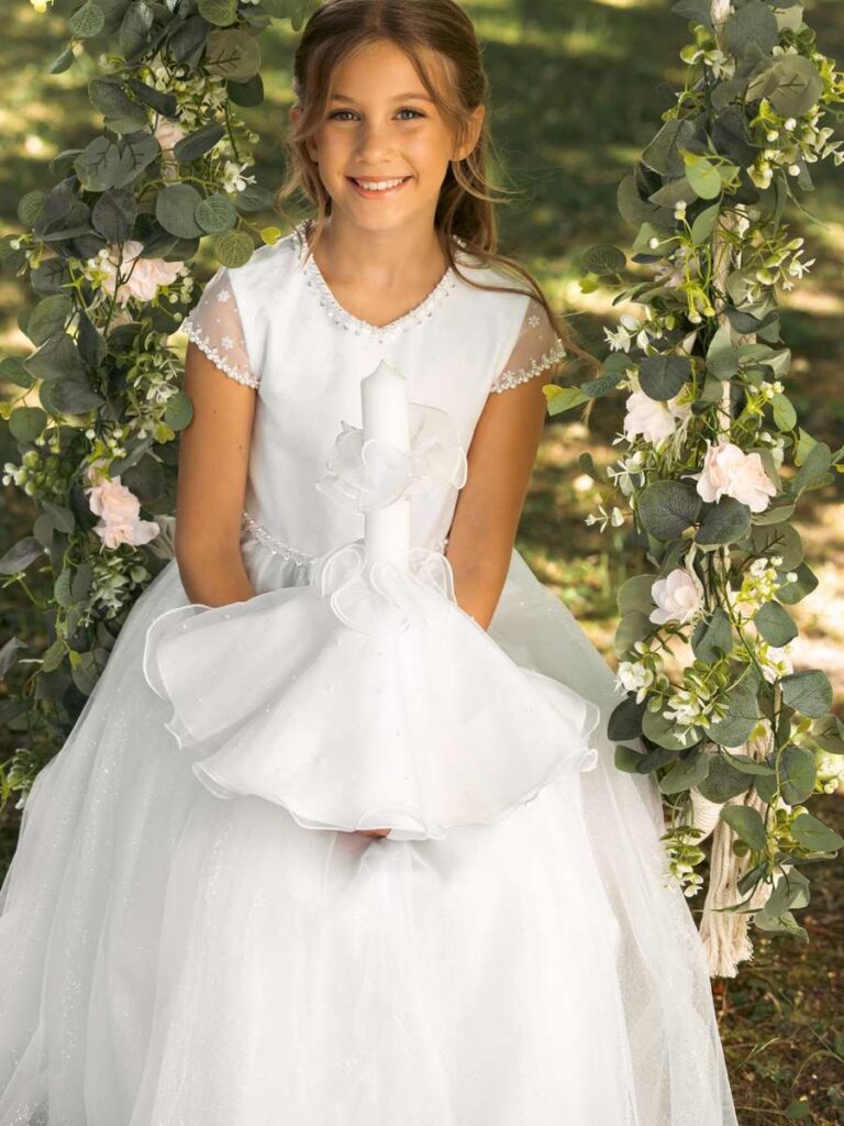 Mädchen im funkelnden Kommunionkleid mit Perlendetails und Kommunionkerze auf einer geschmückten Schaukel – Bianca's Brautmoden Bad Kreuznach