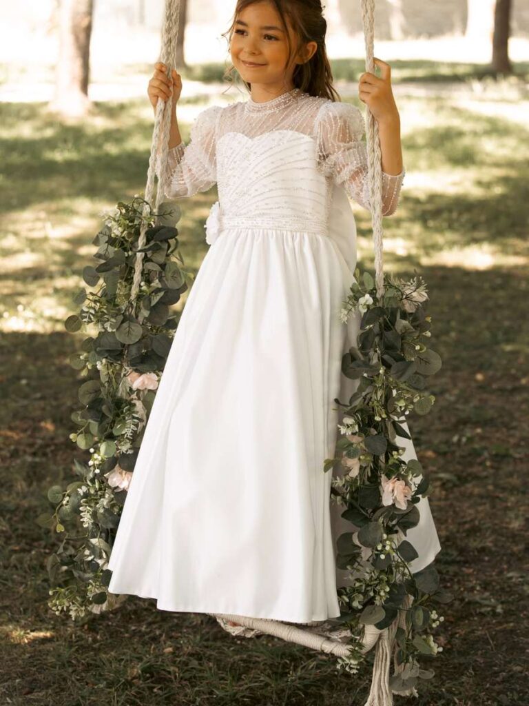 Edles Kommunionkleid mit langen transparenten Ärmeln und Perlenstickerei auf einer Blumenschaukel – Bianca's Brautmoden Bad Kreuznach