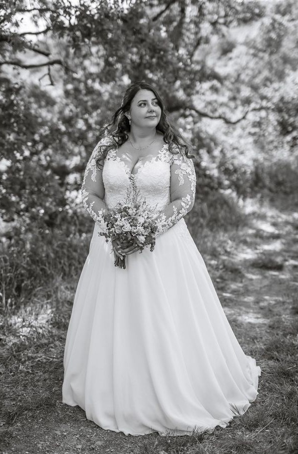 Curvy Braut im Brautkleid mit langen Spitzenärmeln und fließendem Chiffonrock bei einem romantischen Brautshooting im Freien – Bianca's Brautmoden Bad Kreuznach