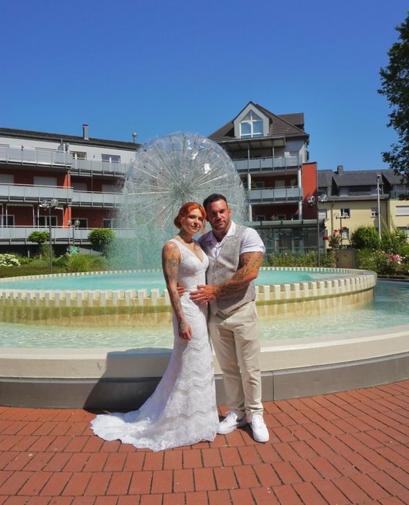 Echtes Brautpaar vor einem Brunnen – Braut im figurbetonten Spitzenbrautkleid und Bräutigam im beigen Hochzeitsanzug von Bianca's Brautmoden Bad Kreuznach