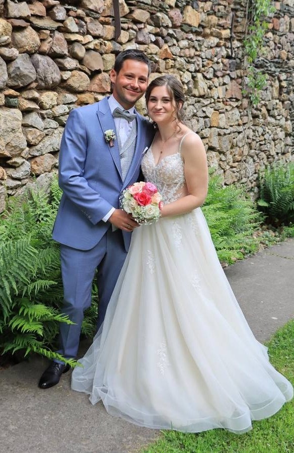 Echtes Brautpaar mit A-Linien Brautkleid und hellblauem Hochzeitsanzug vor einer Natursteinmauer – Bianca's Brautmoden Bad Kreuznach