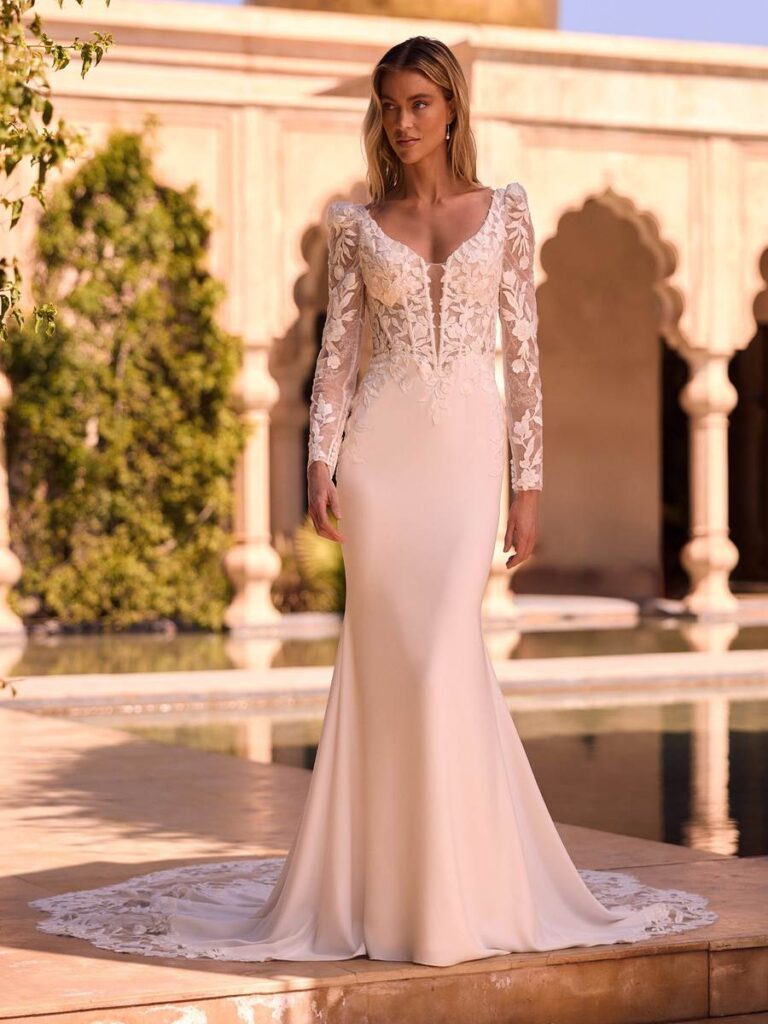 Elegantes Meerjungfrauen-Brautkleid mit cleanem Crepe-Rock, tiefem V-Ausschnitt, langen floralen Spitzenärmeln und romantischer Spitzenschleppe. Das figurbetonte Hochzeitskleid vereint moderne Eleganz mit femininen Details.