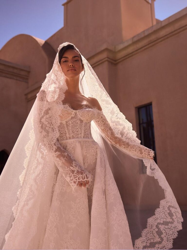 Curvy Fit-and-Flare-Brautkleid mit transparenter Spitzen-Corsage, Off-Shoulder-Ausschnitt, langen Spitzenärmeln und passendem langen Spitzenschleier. Das romantische Hochzeitskleid vereint Eleganz und moderne Raffinesse.