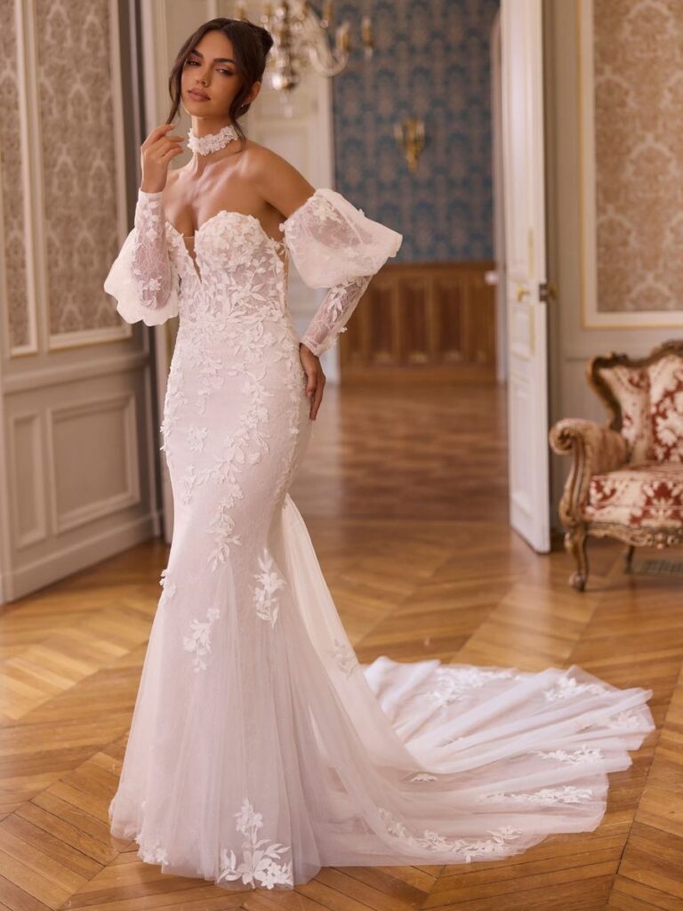 Figurbetontes Meerjungfrauen-Brautkleid mit floraler 3D-Spitze, transparenter Corsage, Off-Shoulder-Puffärmeln und Choker. Das elegante Hochzeitskleid endet in einer romantischen Tüllschleppe.