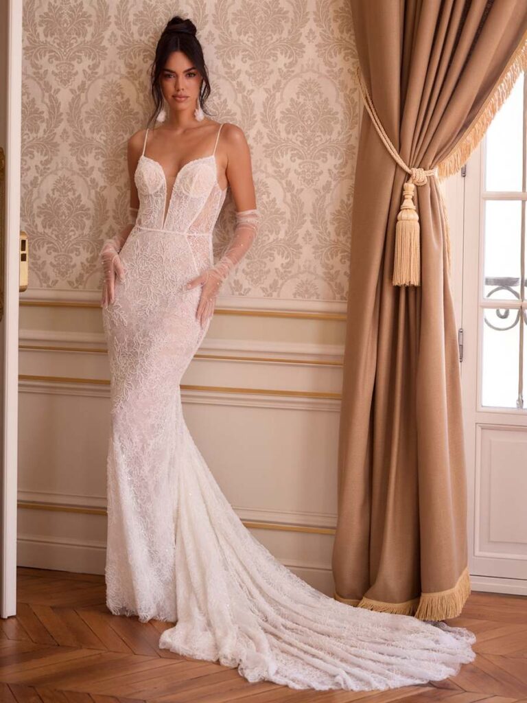 Figurbetontes Meerjungfrauen-Brautkleid aus Spitze mit tiefem V-Ausschnitt, schmalen Spaghettiträgern, transparenter Corsage und langen Tüll-Armstulpen. Das elegante Hochzeitskleid endet in einer romantischen Spitzenschleppe.